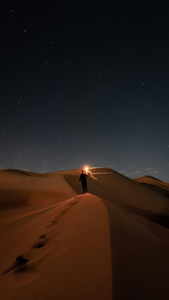 A lone figure walks across desert dunes beneath a starry night sky, the light guiding their way. Perfect for nature and adventure themes.